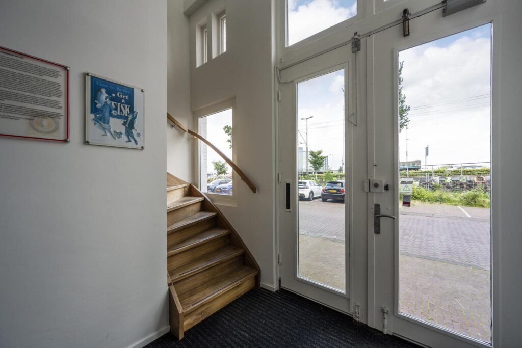 Ingang met houten trap en glazen voordeuren aan de De Ruijterkade, uitkijkend op een parkeerplaats.